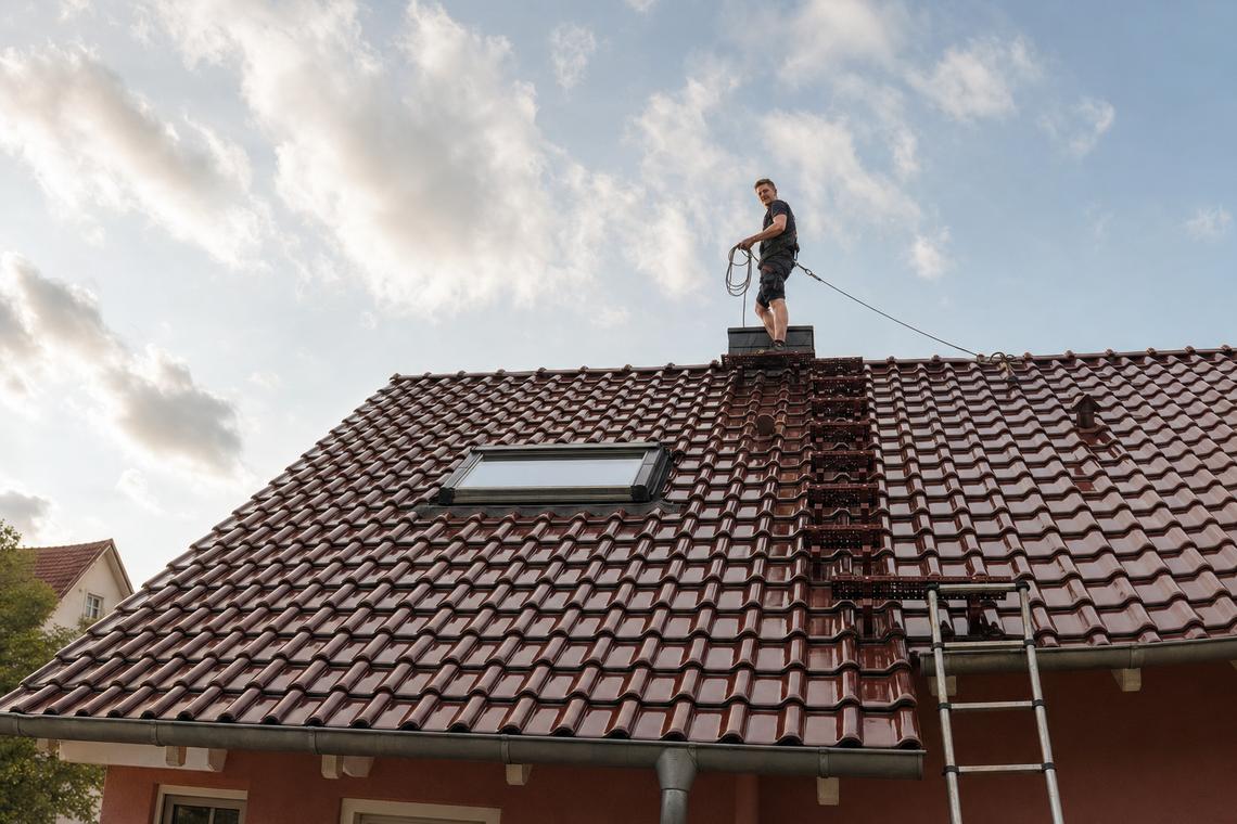 Ein Schornsteinfeger von haushoch4 arbeitet am Schornstein auf einem Ziegeldach. Eine Leiter lehnt an der Dachkante, darüber Himmel mit Wolken.